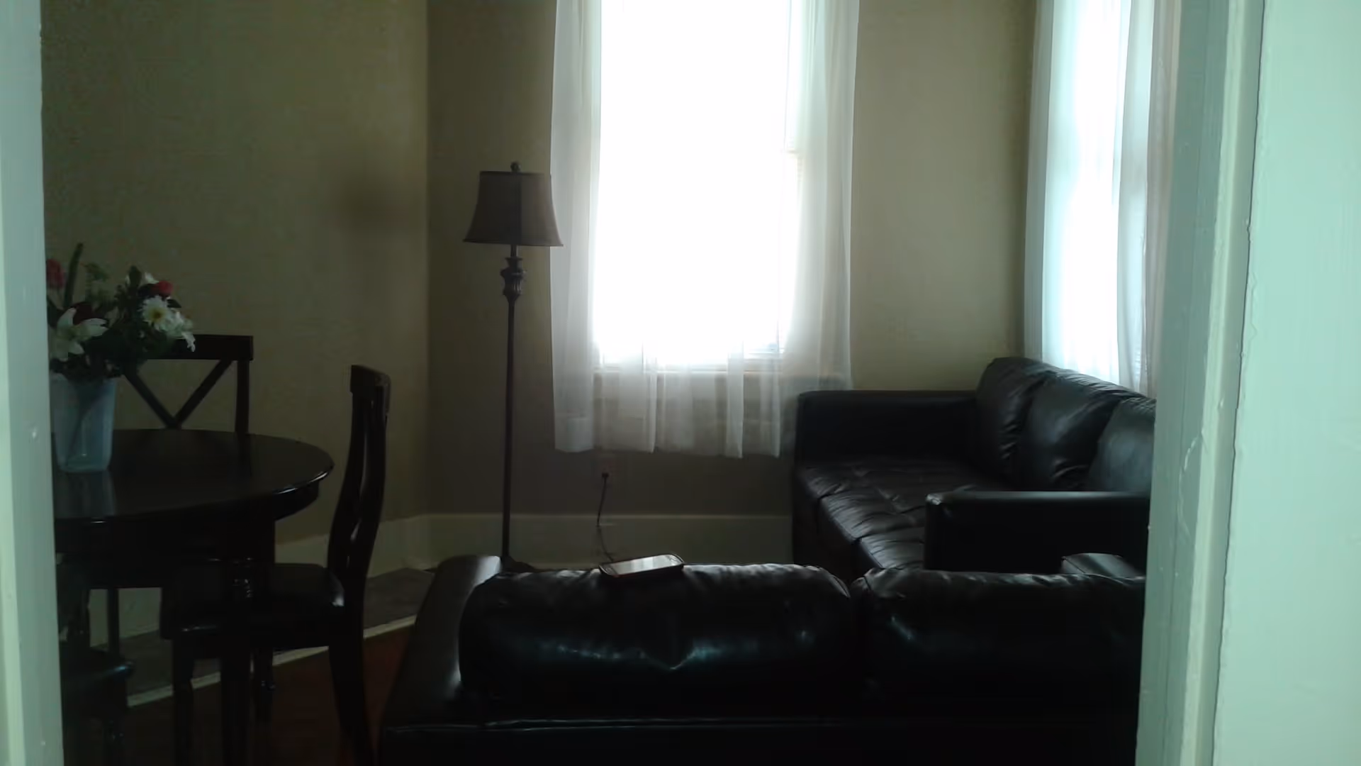 Small living room with black leather sofas, a floor lamp by a curtained window, and a round dining table with chairs and a vase of flowers.