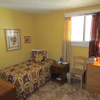 A small bedroom with a single bed covered in a patterned bedspread featuring maroon and yellow colors. There is a wooden nightstand next to the bed with a bottle and some items on it. A wooden chair is placed near a window with partially drawn curtains. A wooden dresser with books and a lamp on top is also visible. The walls are painted yellow and there are two framed pictures hanging on the wall.