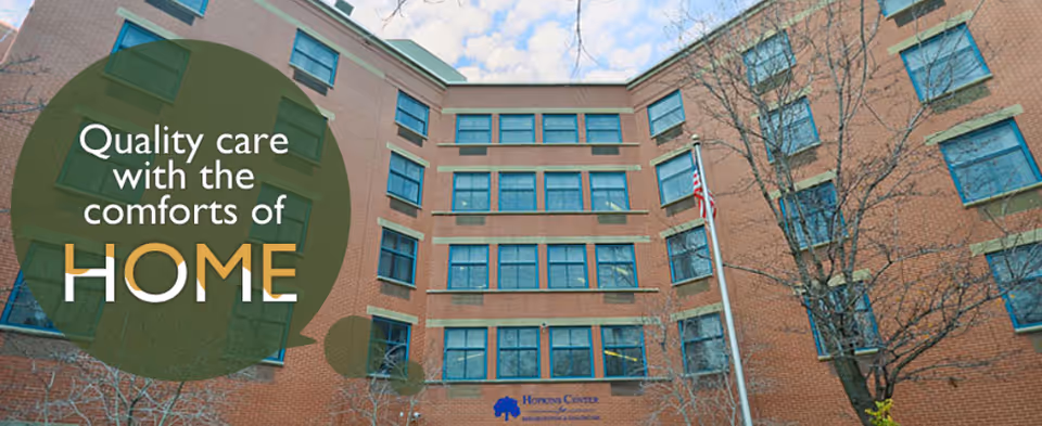 Exterior brick facility with American flag, trees, and slogan 'Quality care with the comforts of HOME'.