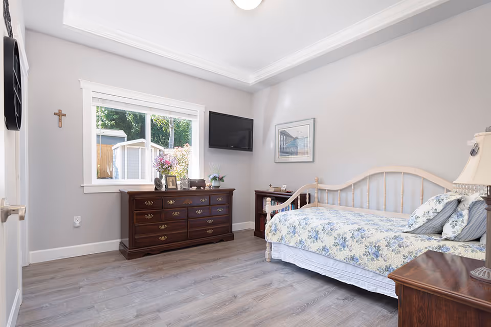 Bright bedroom with a white bed, floral quilt, dark dresser, window, TV, and cross.