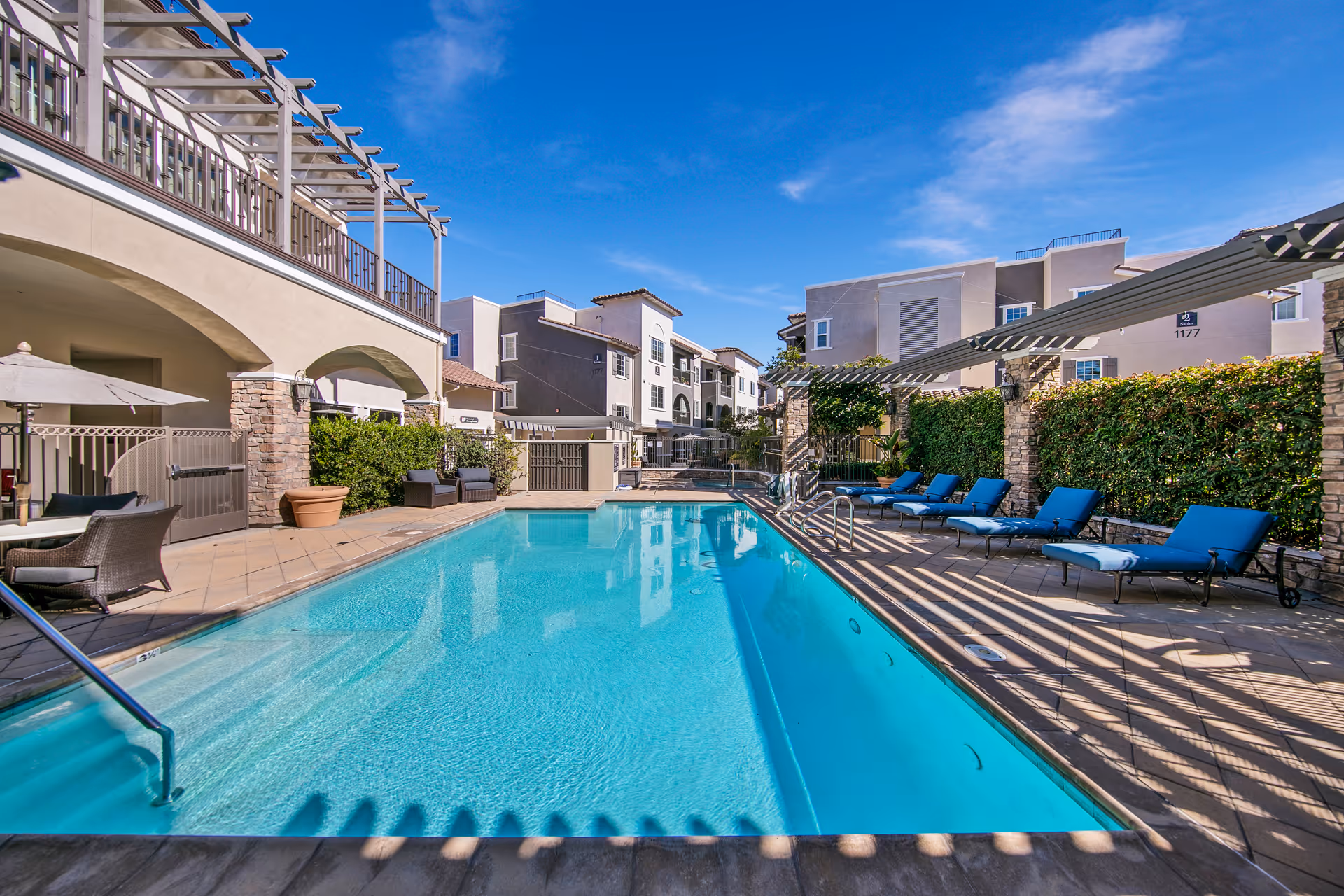 Outdoor pool area with blue lounge chairs, umbrellas, and surrounding buildings.