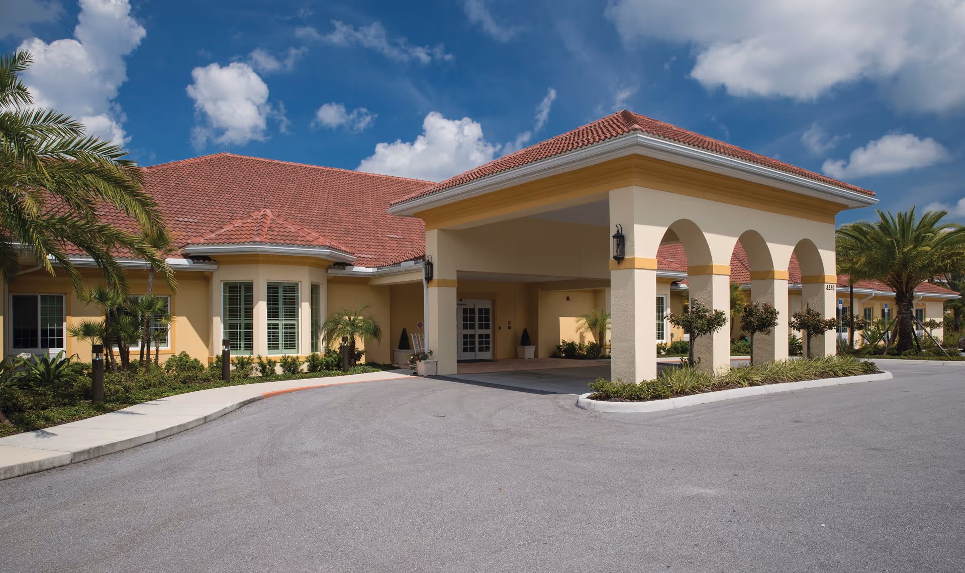 Exterior view of Windsor Reflections Memory Care Of Lakewood Ranch showing a building with a red tiled roof, beige walls, arched entrance, palm trees, and a clear blue sky with some clouds.