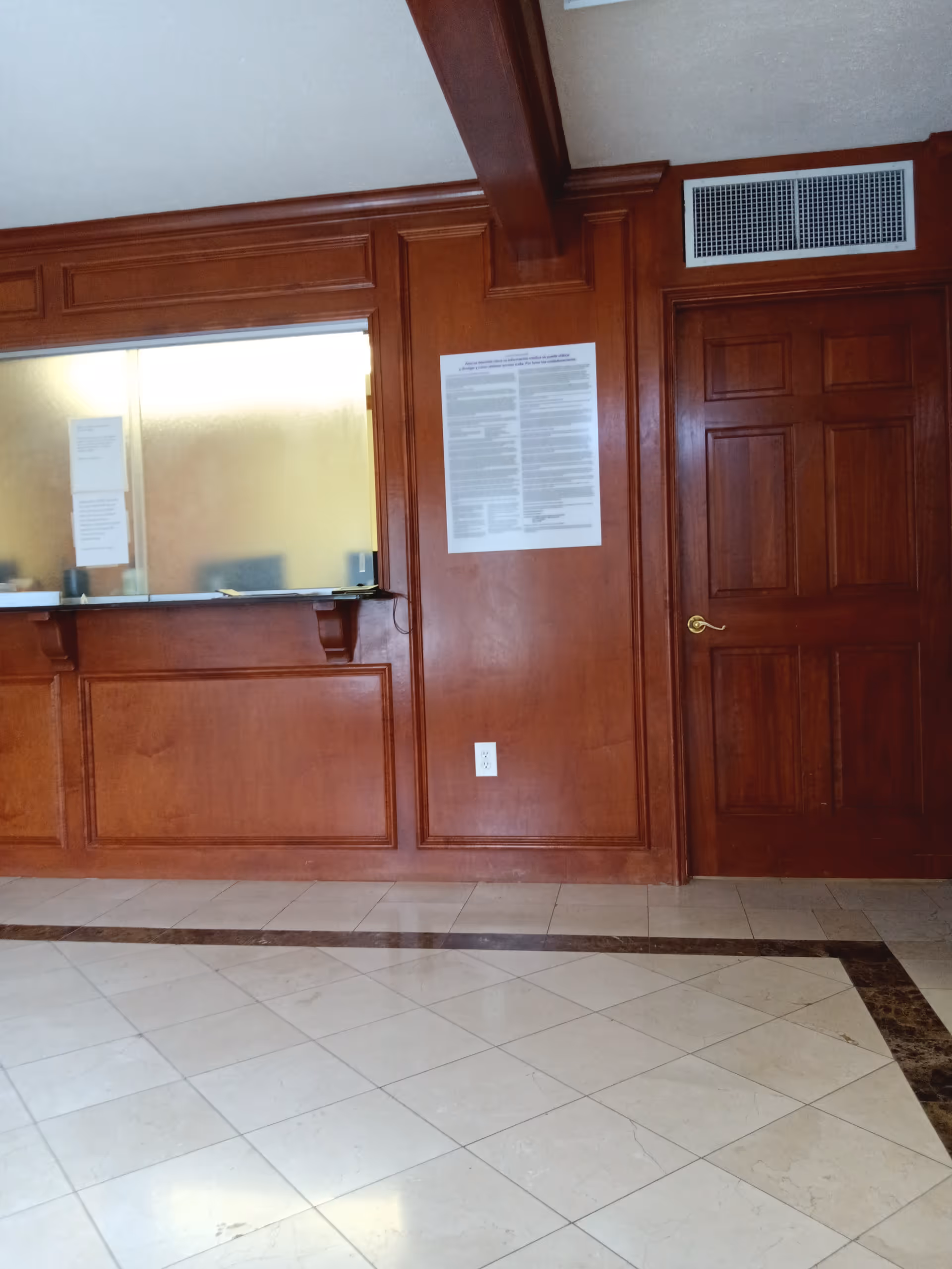 Wood-paneled lobby with a reception window, posted notices, and a closed wooden door.
