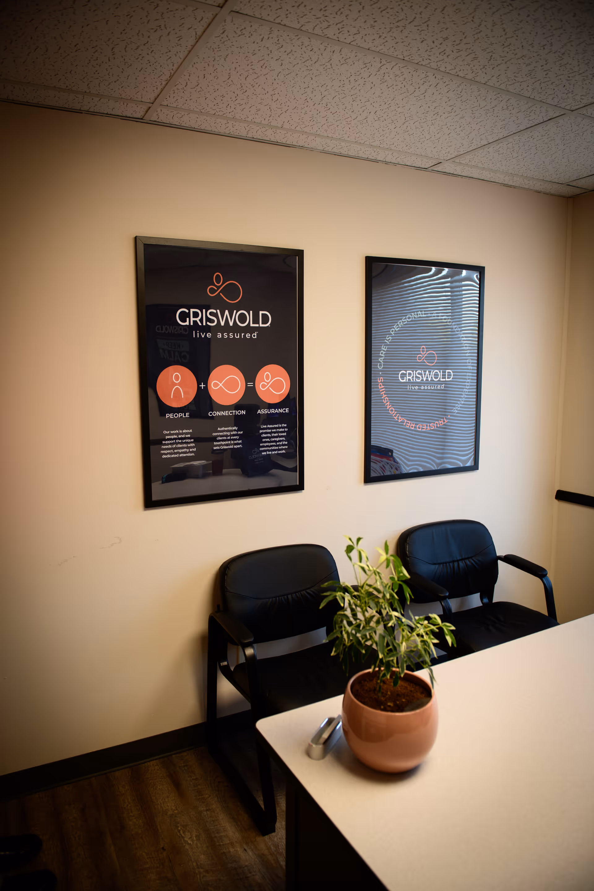Two Griswold posters hang on beige wall beside black chairs and a plant on a desk.