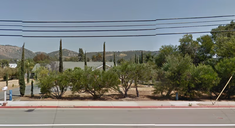 Busy street view with trees and shrubs along a sidewalk, hills in distance under clear sky.