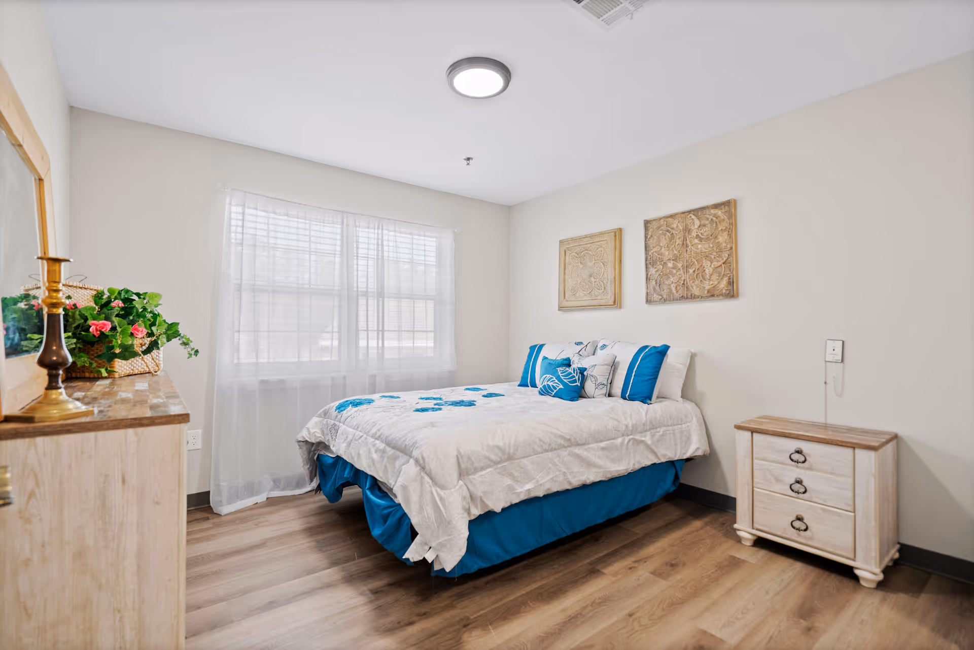 A tidy bedroom in a senior living facility with a bed, dresser, and window.