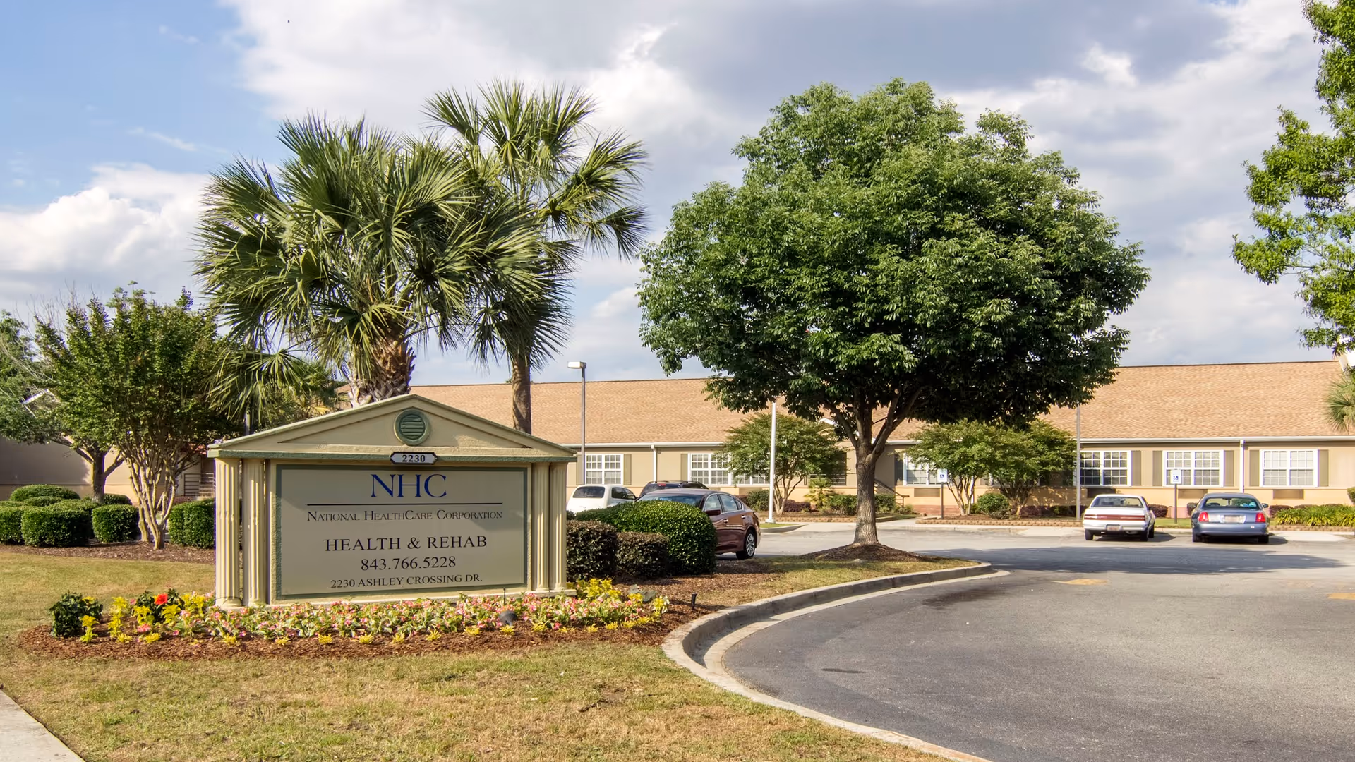 Exterior view of NHC Health Care Charleston with sign, palm trees, and parked cars.