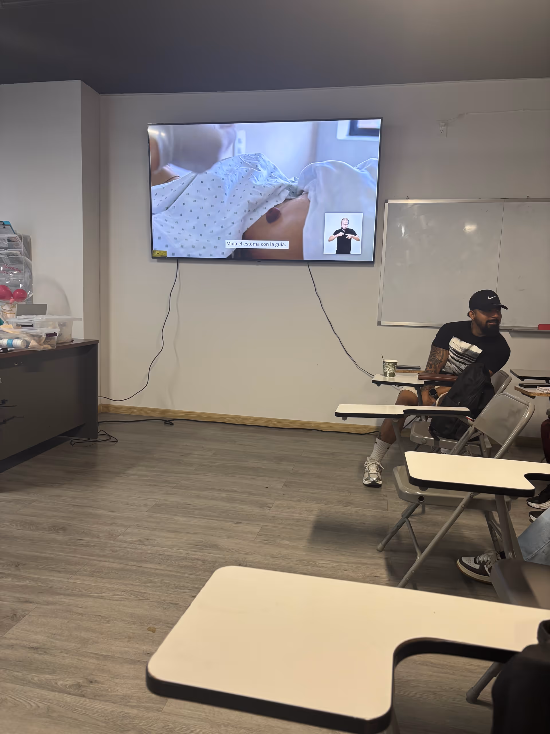 A classroom-style training room with a wall-mounted TV and a man seated at a desk.