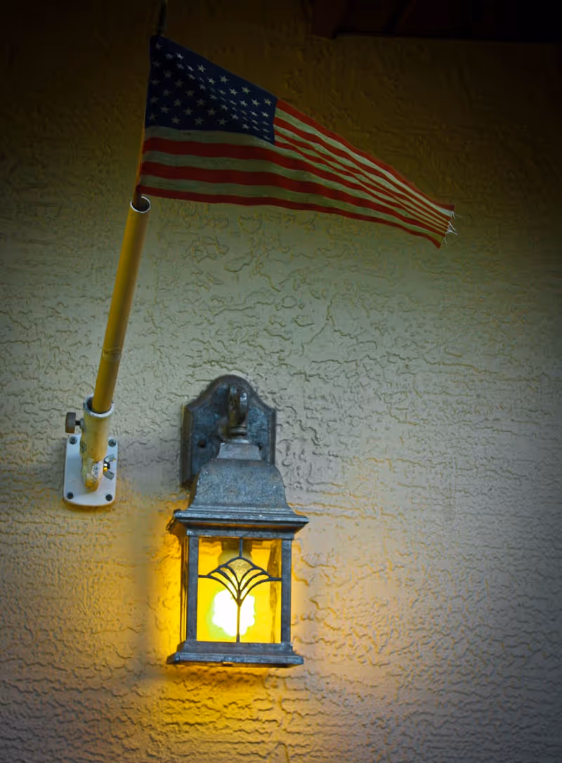 A lit outdoor wall lantern mounted on a textured stucco wall beneath a small American flag.