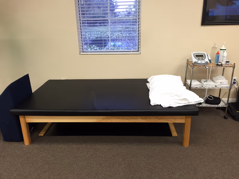 A padded therapy table with pillow in a small clinic room, a medical cart nearby.