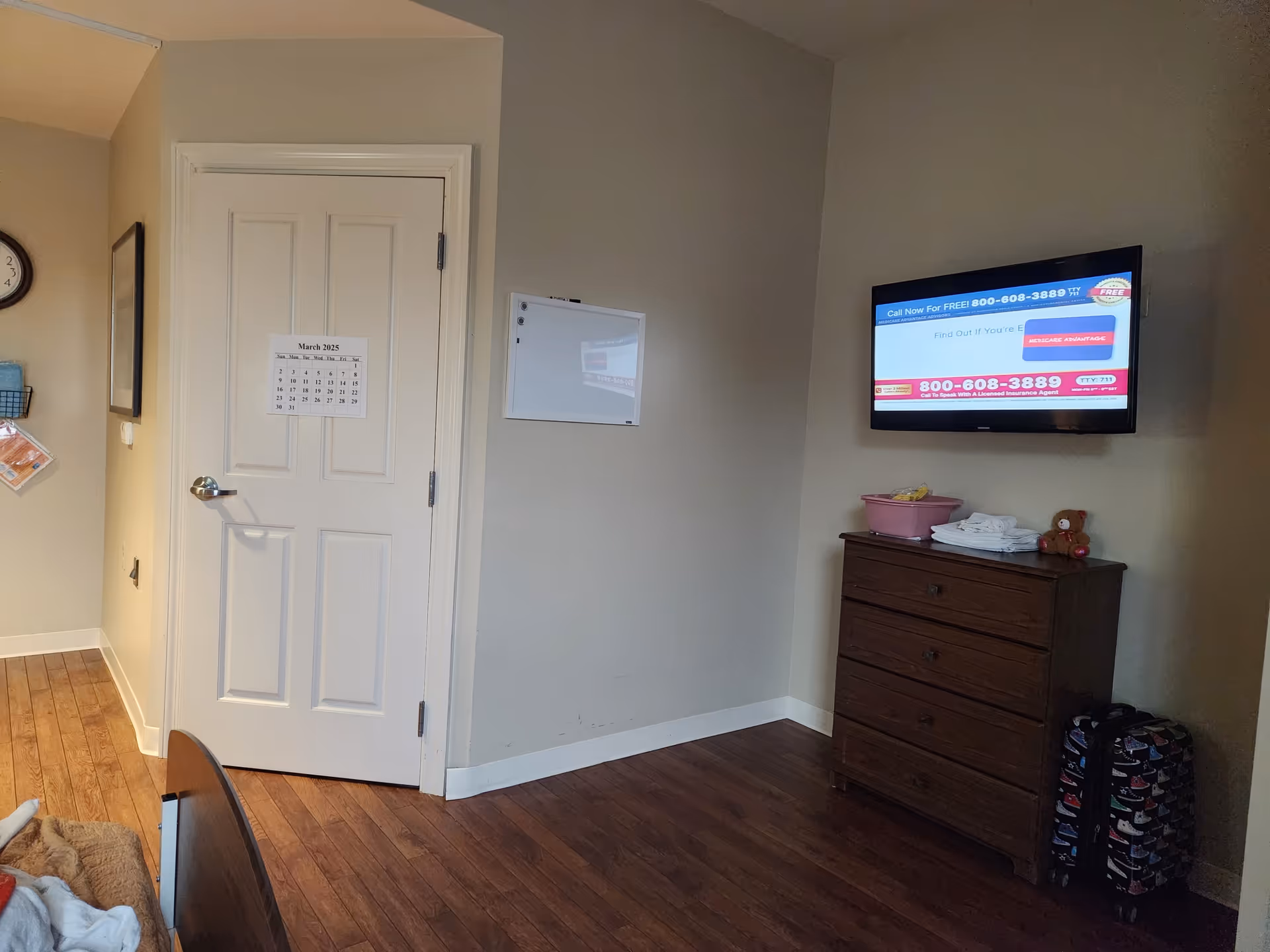 Interior of a resident room with door, calendar, TV, dresser, and suitcase.