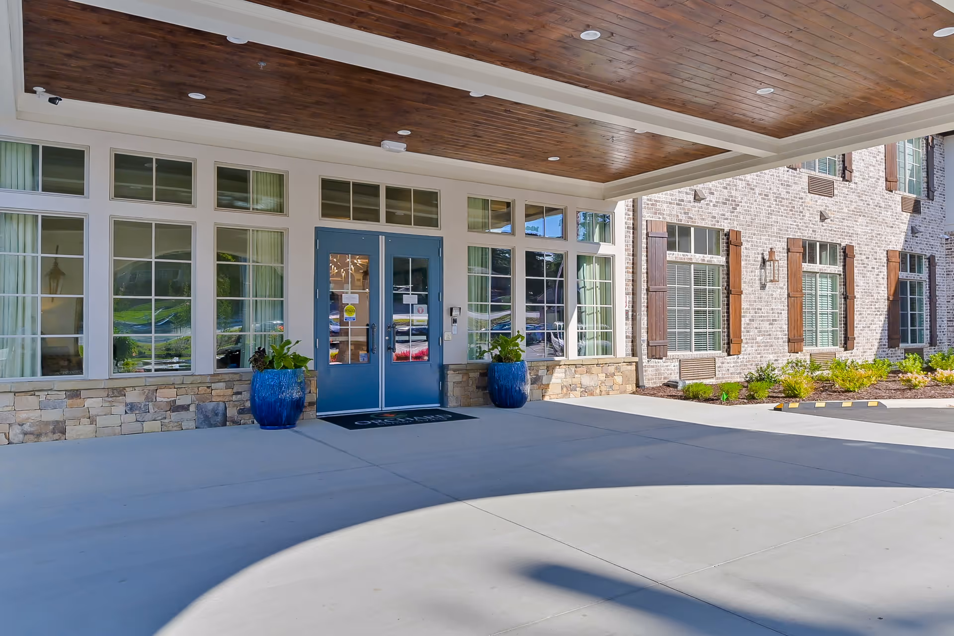 Covered main entrance of a senior living building with double blue doors, large windows, stone and brick facade, and potted plants.