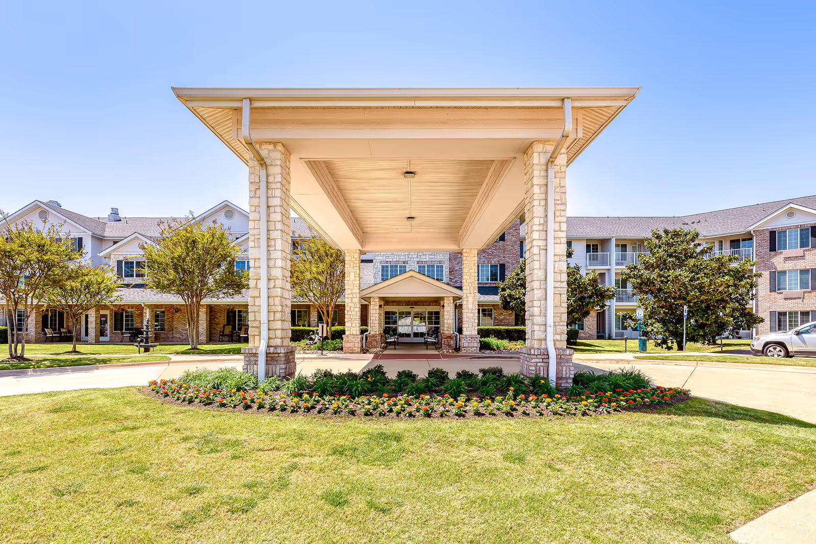 Front entrance of a multi-story senior living building with a covered porte-cochere, landscaped lawn, and flowerbeds.