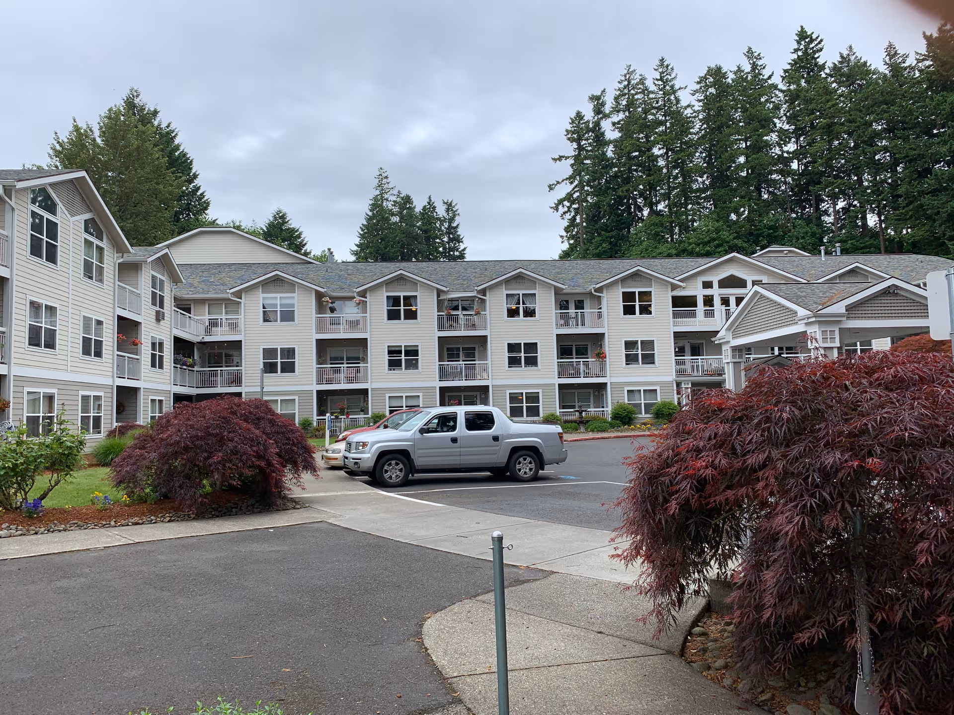 Three-story beige senior apartment building with balconies, landscaping, and a silver pickup in parking lot.