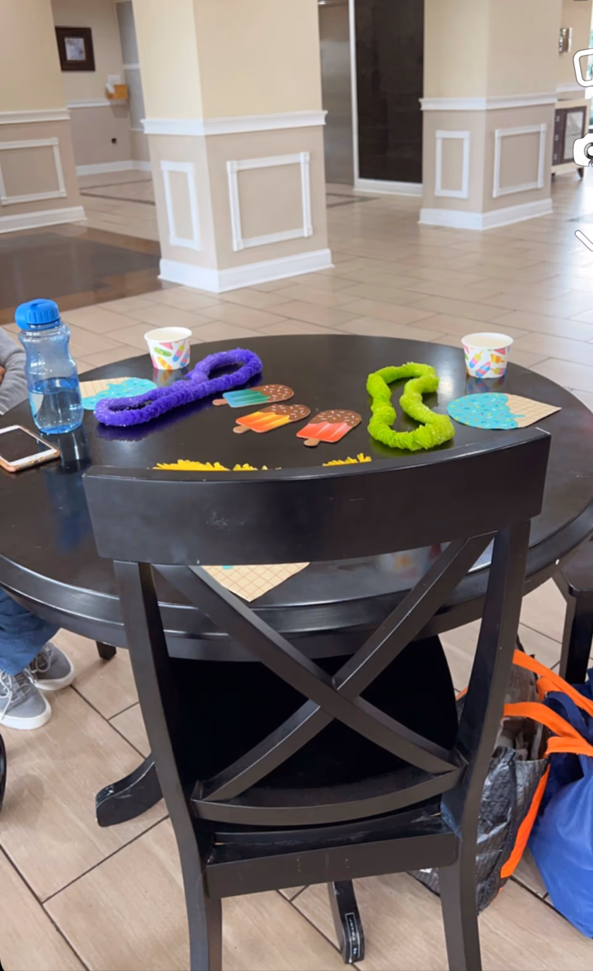 Round black table with crafts, cups, and water bottle in a senior living facility lobby.