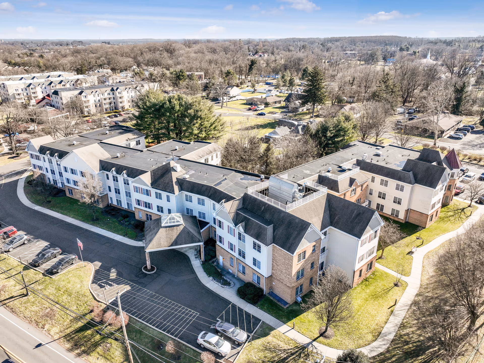 Aerial view of a large senior living facility building with multiple wings, surrounded by trees and parking lots. The building has a mix of brick and white siding with a covered entrance. The surrounding area includes other residential buildings and open green spaces under a clear sky.