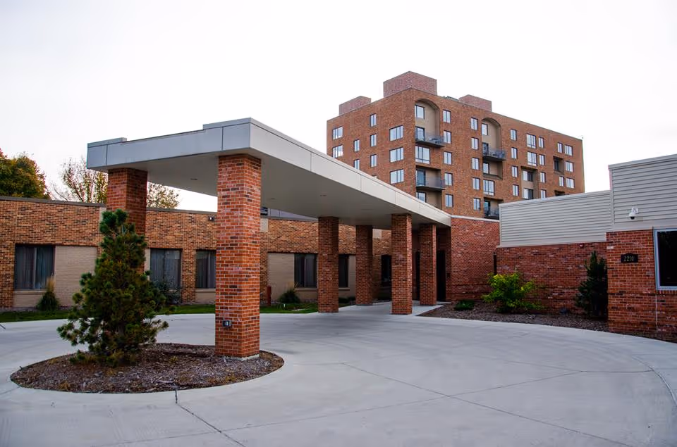 Covered brick entrance and porte-cochere of a multi-story brick senior living facility with a circular driveway and small landscaped islands.