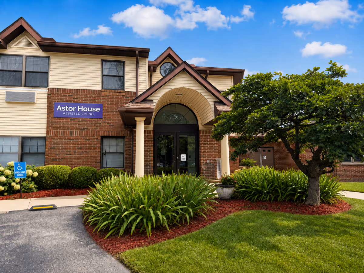 Exterior front of Astor House Assisted Living with brick facade, arched entry, and lush landscaping.