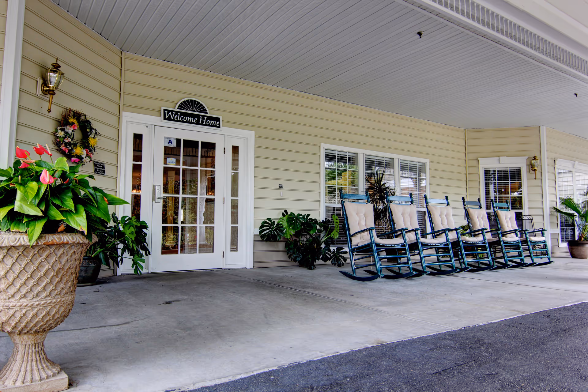 Covered entrance area of a senior living facility with a row of cushioned rocking chairs, potted plants, and a glass door with a 'Welcome Home' sign above it.