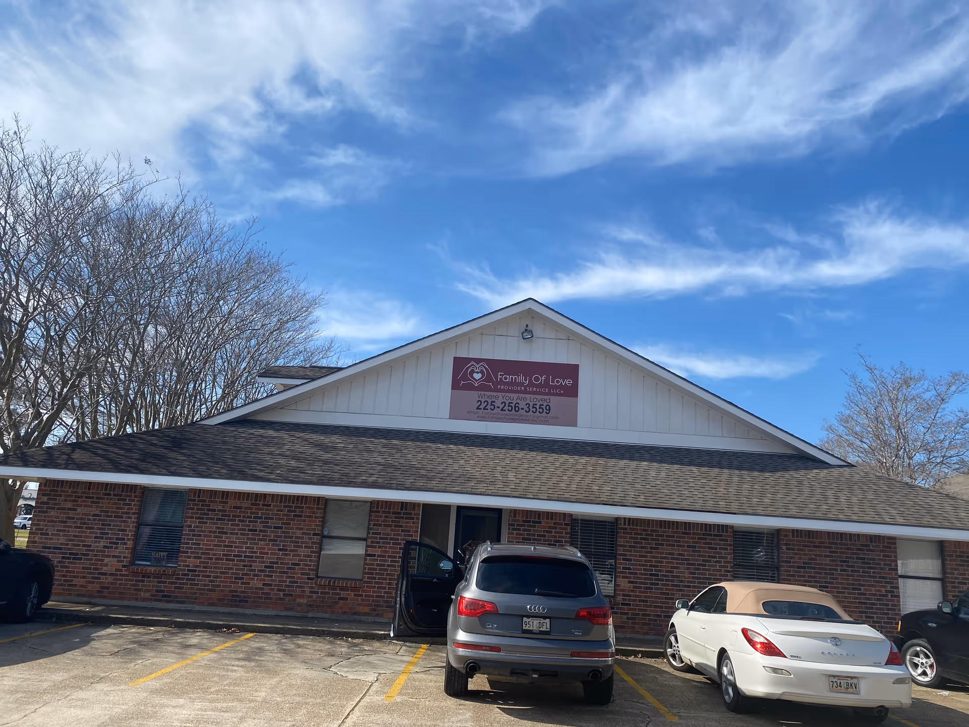 Front view of a brick facility with a Family Of Love sign and parked cars.