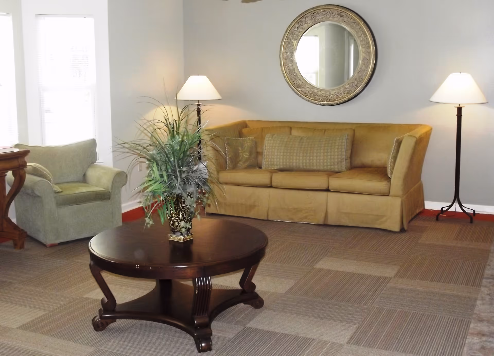 A living room with a tan sofa adorned with three cushions, a green armchair, two floor lamps with white shades, a round dark wooden coffee table with a decorative plant in a vase, and a large ornate round mirror hanging on a gray wall.