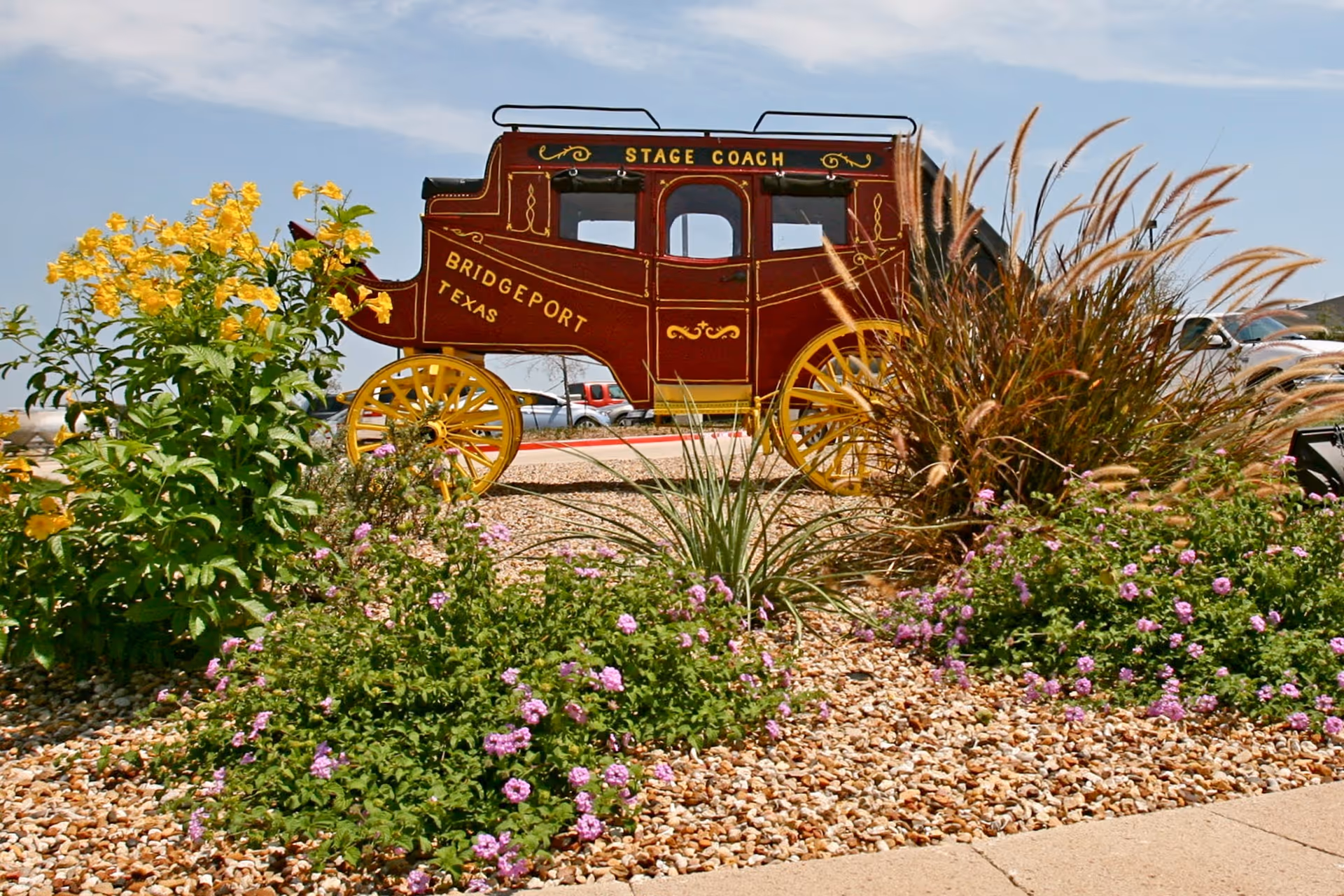 Red vintage stagecoach sculpture with yellow wheels among rocks and flowering plants.
