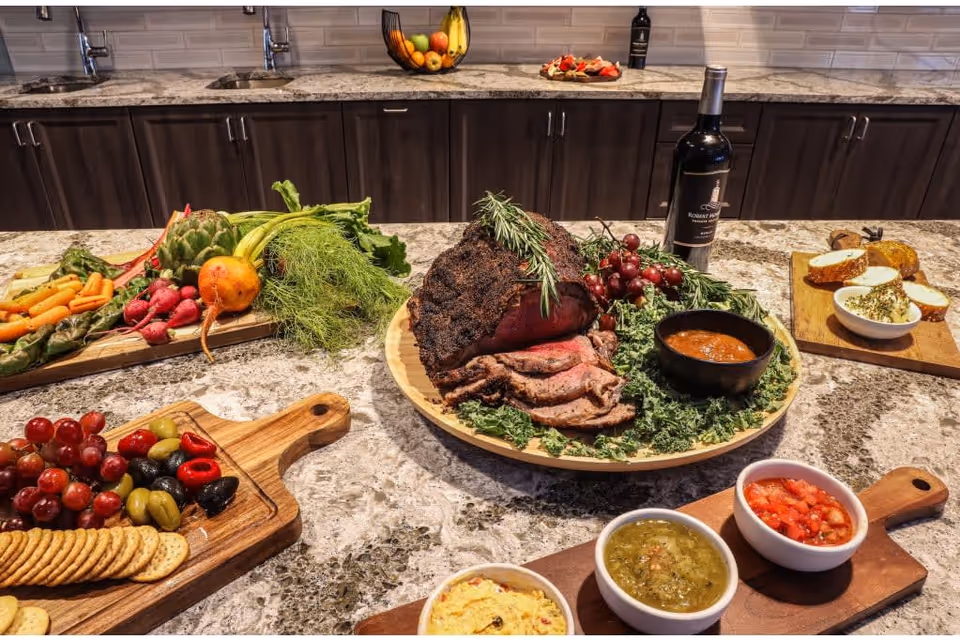 A kitchen countertop with an assortment of food including sliced roast beef garnished with rosemary and grapes, a bottle of red wine, fresh vegetables on a cutting board, crackers with olives and grapes, and several bowls of dips and sauces. The background shows a kitchen sink and dark wooden cabinets with a tiled backsplash.