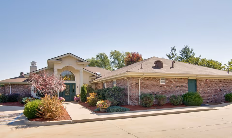 Exterior view of a single-story brick building with a green door entrance, surrounded by landscaped bushes and small trees under a clear blue sky.