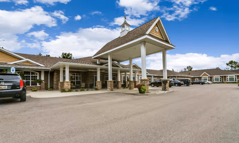 Exterior view of Candlestone Assisted Living & Memory Care facility showing a covered entrance with white pillars and stone bases, multiple parked cars, and a clear blue sky with some clouds.