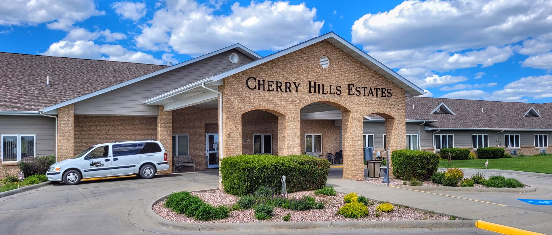 Exterior view of Cherry Hills Estates Assisted Living & Independent Apartments building with a covered entrance, a white van parked on the driveway, and landscaped bushes and plants under a partly cloudy blue sky.