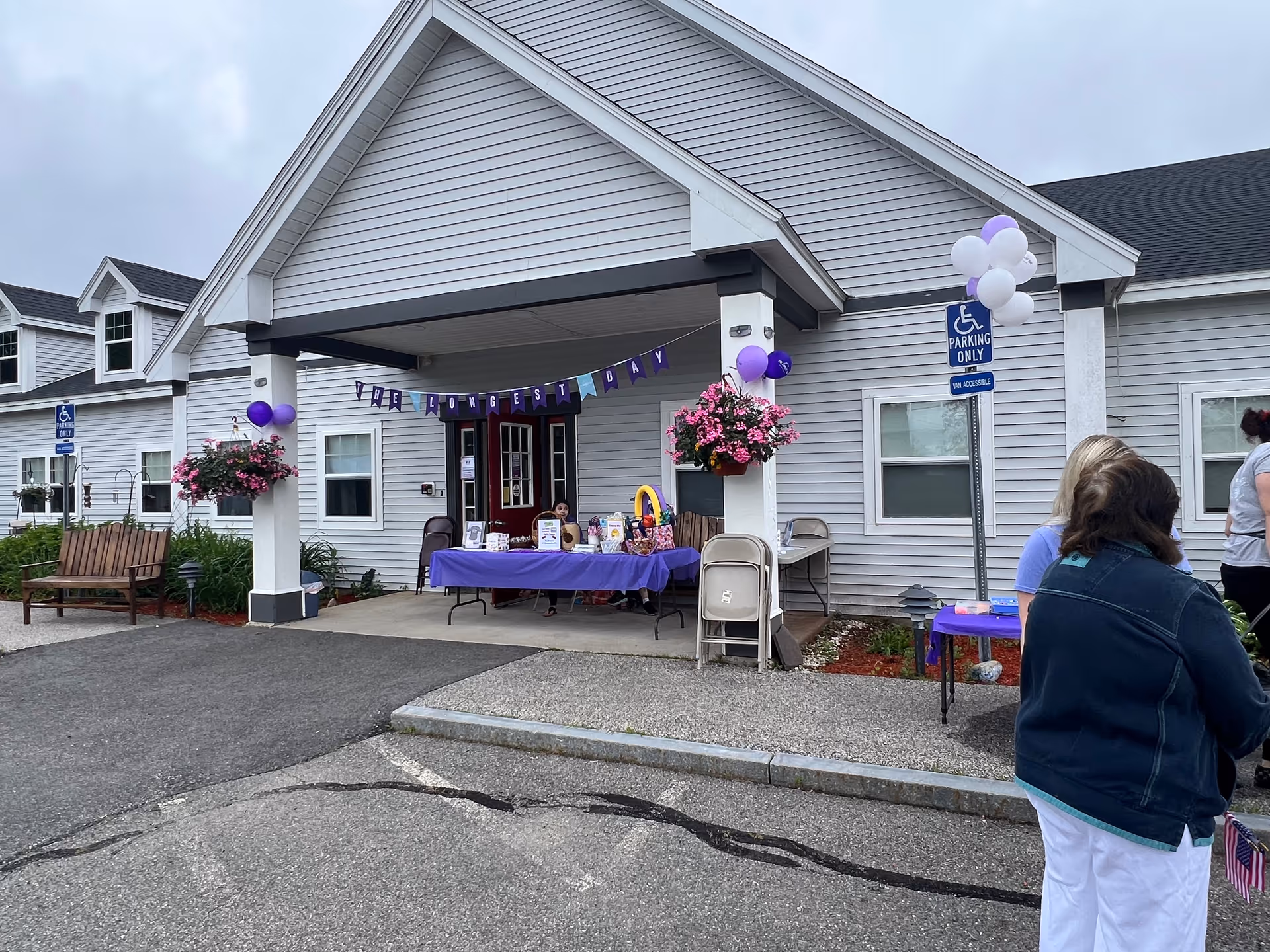 Entrance of a senior living facility with a covered porch decorated with purple and white balloons and a banner that reads 'THE LONGEST DAY'. There are tables set up with informational materials and people standing nearby. The building is light gray with white trim and has a handicap parking sign visible.