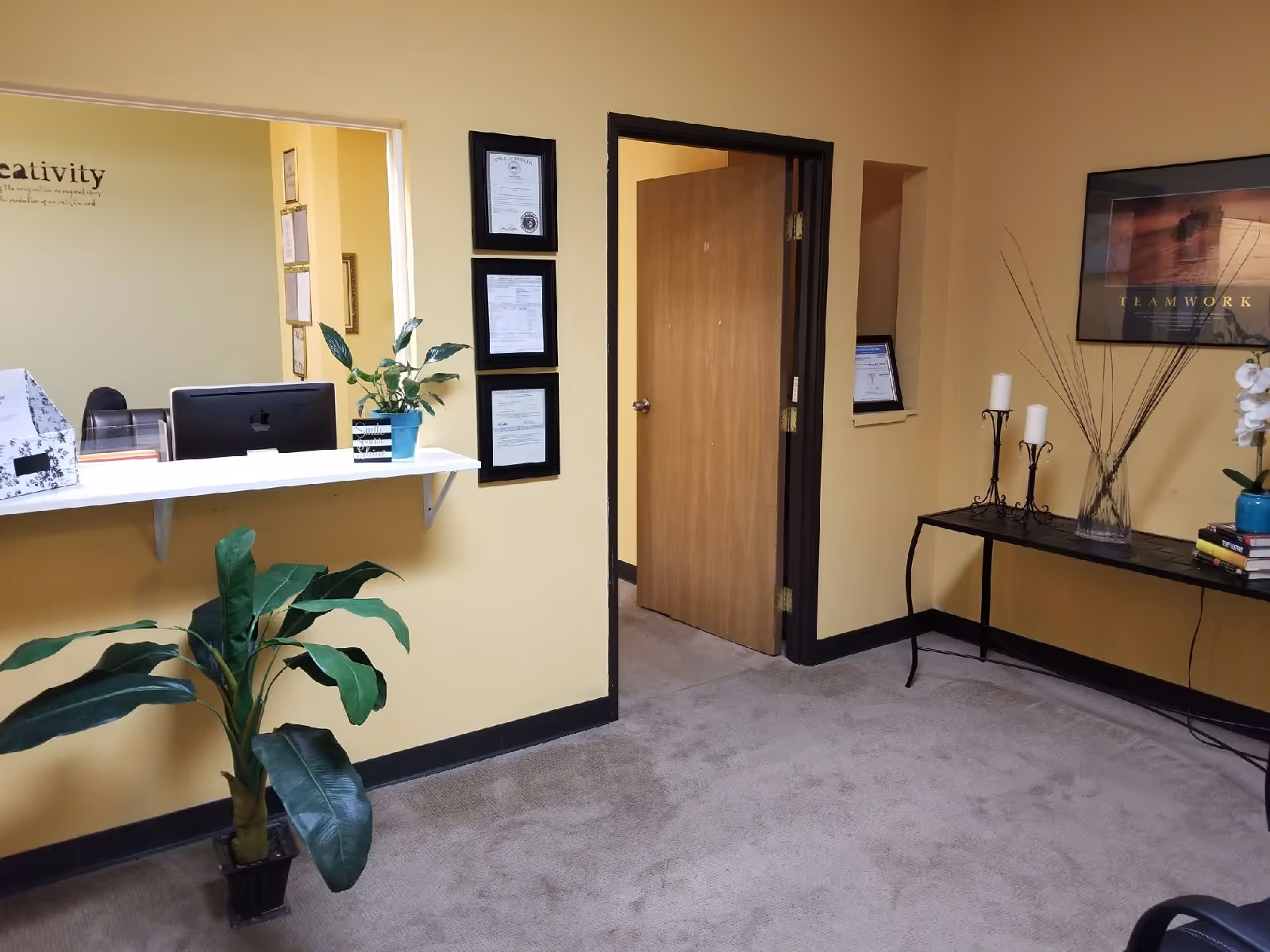 Reception area in a senior living facility with a desk, computer, framed certificates, and plants.