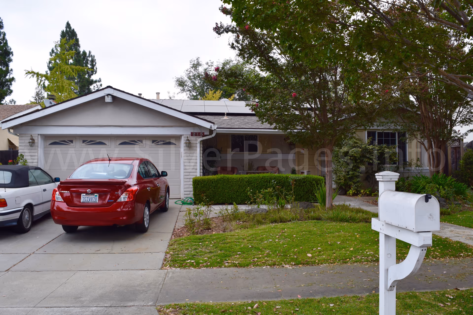 A single-story residential front view with a driveway, cars, and greenery.