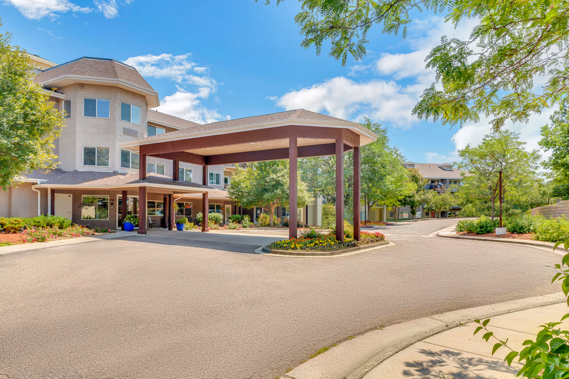 Entrance area of Quincy Place | Sky Active Living facility with a covered drop-off zone, surrounded by trees and landscaping under a blue sky with scattered clouds.