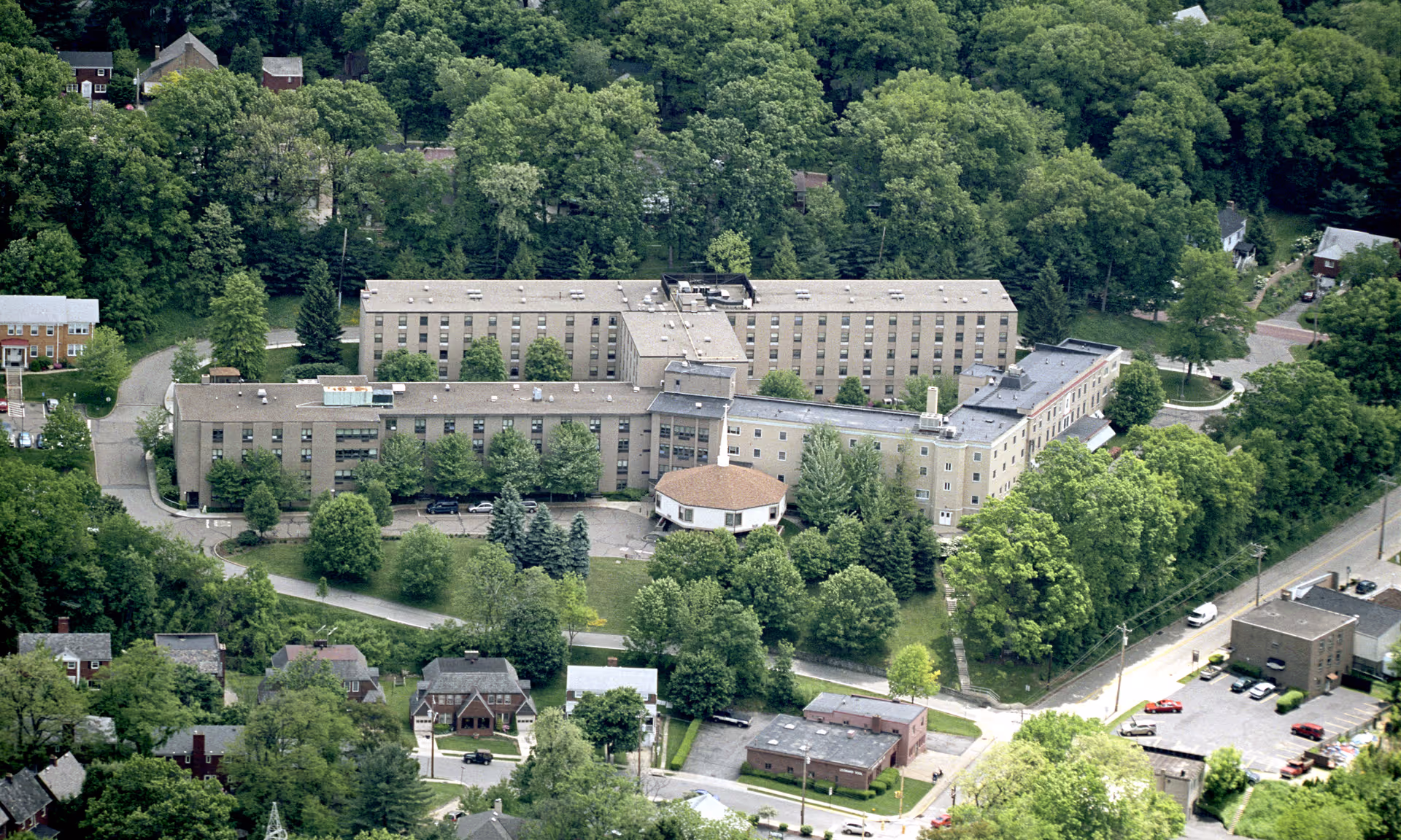 Aerial view of a large senior living facility named Baptist Homes surrounded by dense green trees and residential houses. The building is multi-storied with a central circular section and several wings extending outward. There are parking areas and roads visible around the facility.