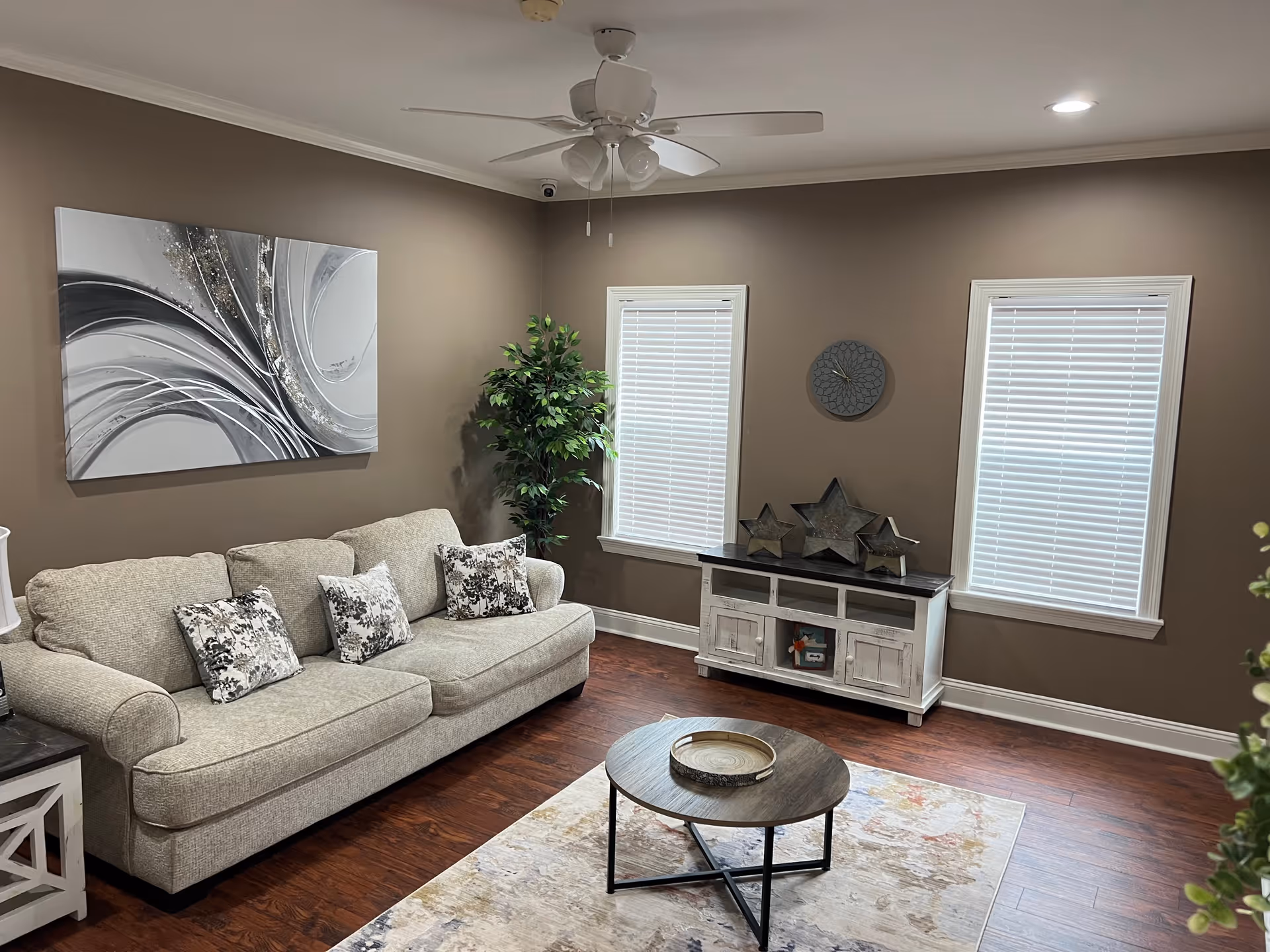 A cozy living room with a beige sofa adorned with three patterned cushions, a round wooden coffee table on a light-colored rug, a white side table with a lamp, two windows with white blinds, a white wooden cabinet with decorative star ornaments, a wall clock, a large abstract painting, a ceiling fan with lights, and a potted plant in the corner.