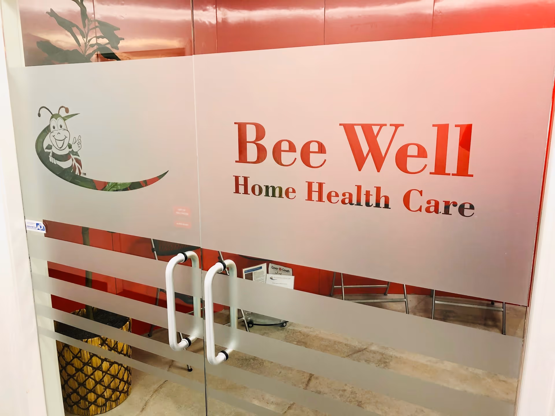 Glass door entry with Bee Well Home Health Care sign and logo, metal handles.