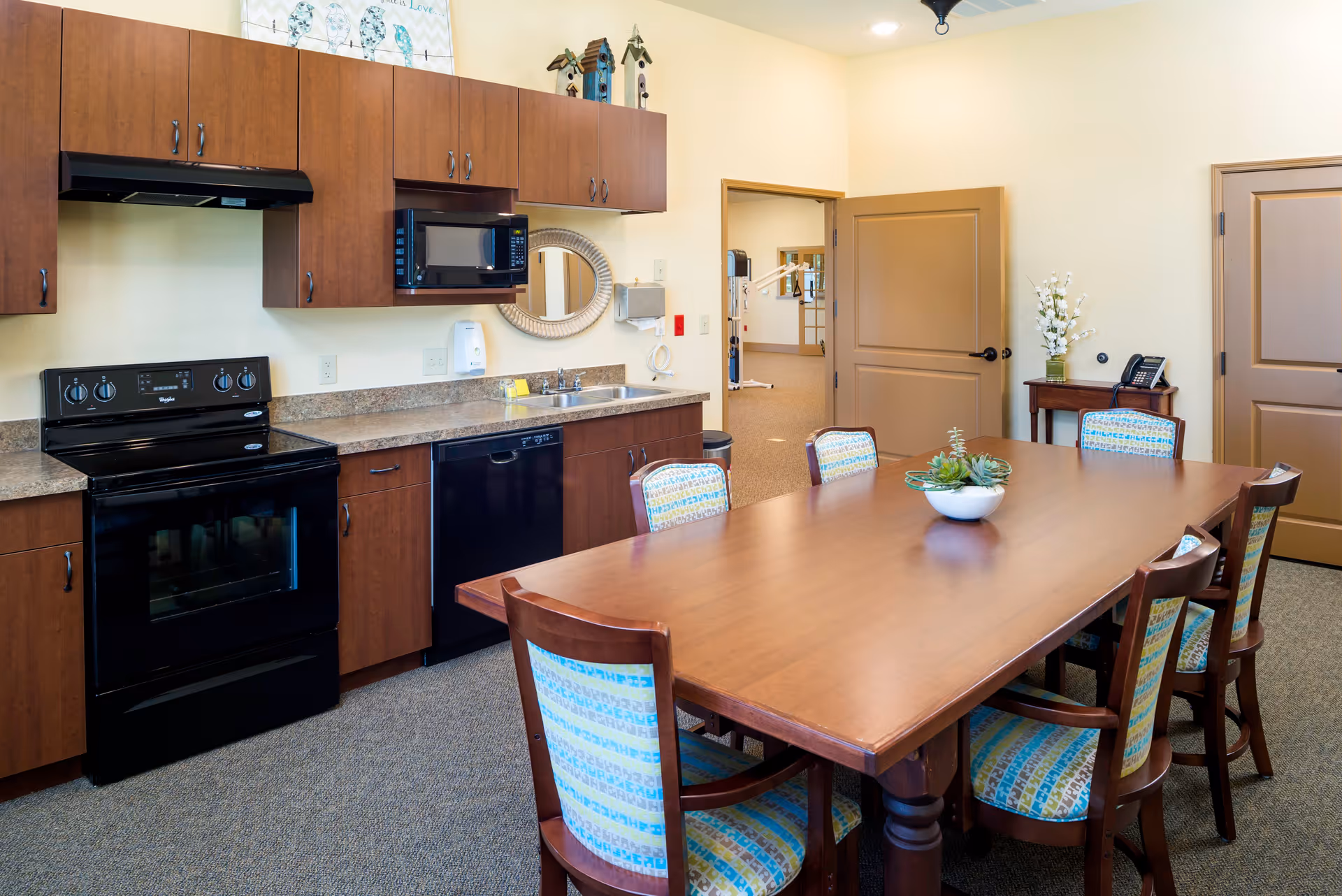 A kitchen and dining area in a senior living facility with wooden cabinets, a black stove, microwave, dishwasher, and a double sink. A wooden dining table with six chairs featuring patterned upholstery is in the foreground, with a small plant centerpiece. The room has beige walls, carpeted flooring, and two closed doors. A small table with a phone and a vase of flowers is against the wall.