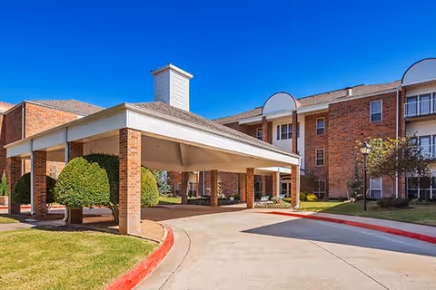 Exterior view of a senior living facility with a covered driveway entrance, brick building, manicured bushes, and clear blue sky.