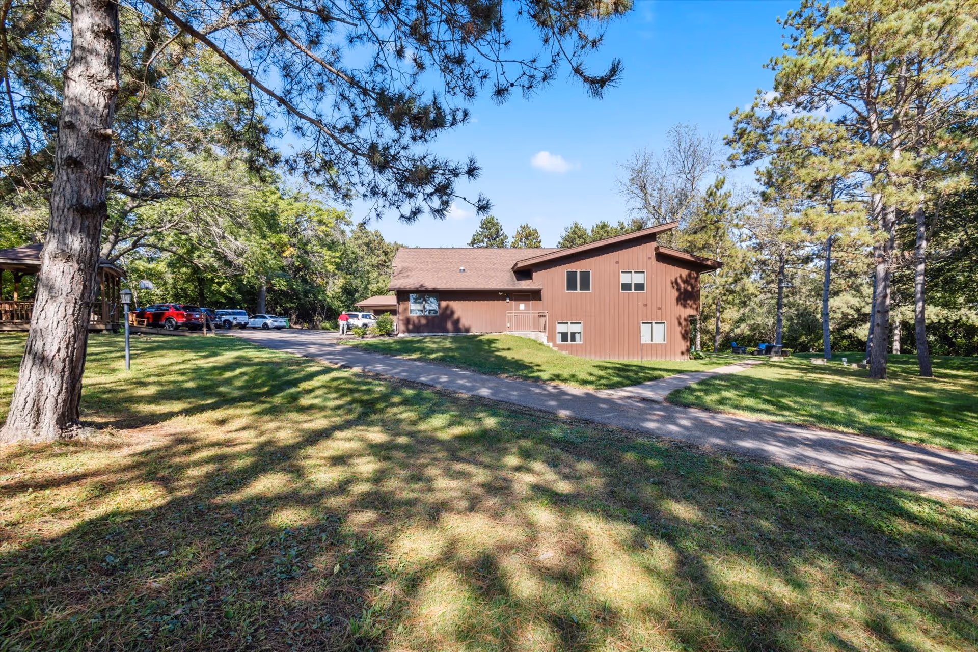 Brown, two-story building surrounded by green lawn and tall trees, with a paved path to entrance.