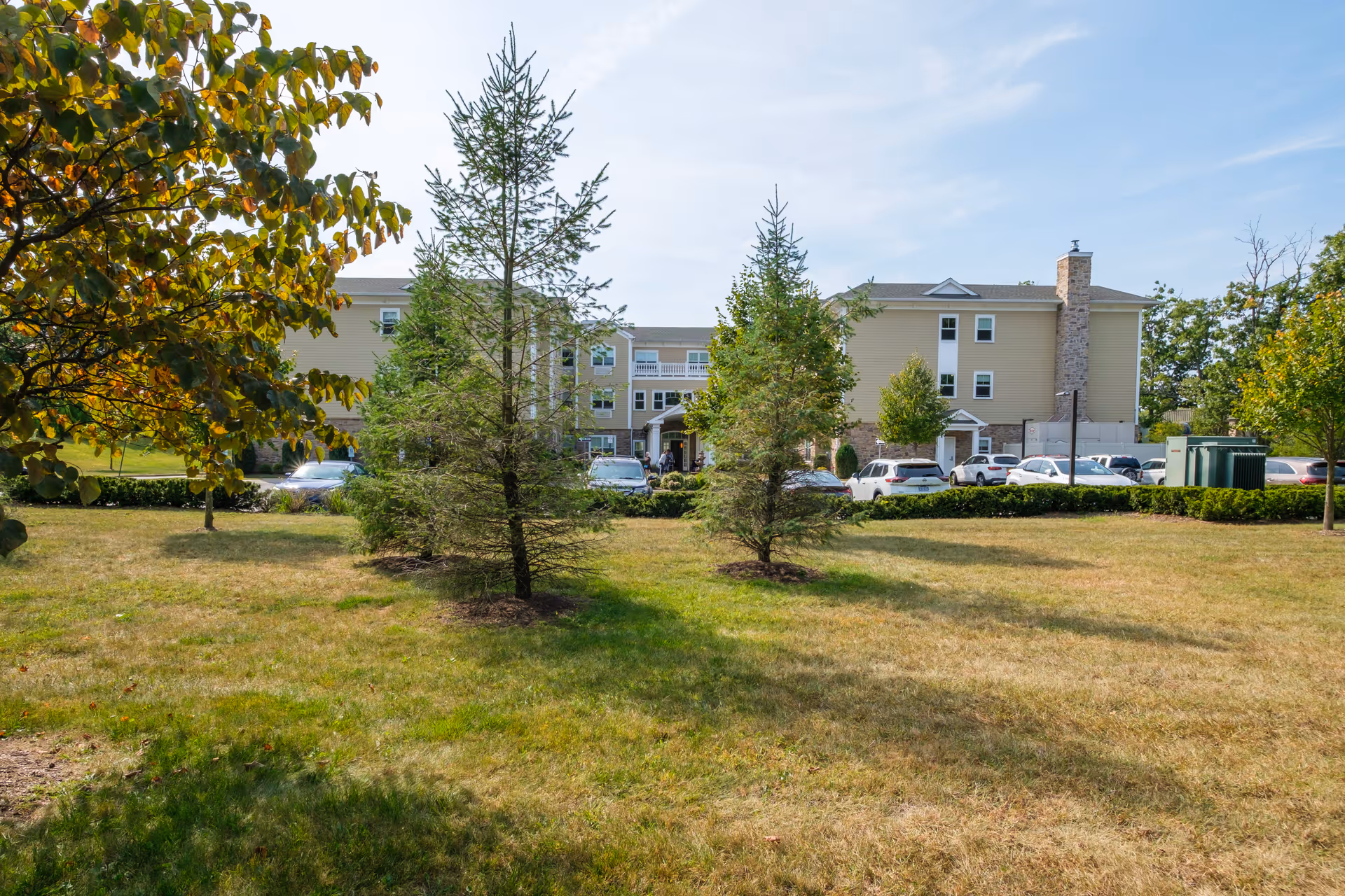 View of a senior living facility building named Amber Court seen from a grassy lawn with several small evergreen trees and other leafy trees. There are parked cars in front of the building under a clear blue sky.