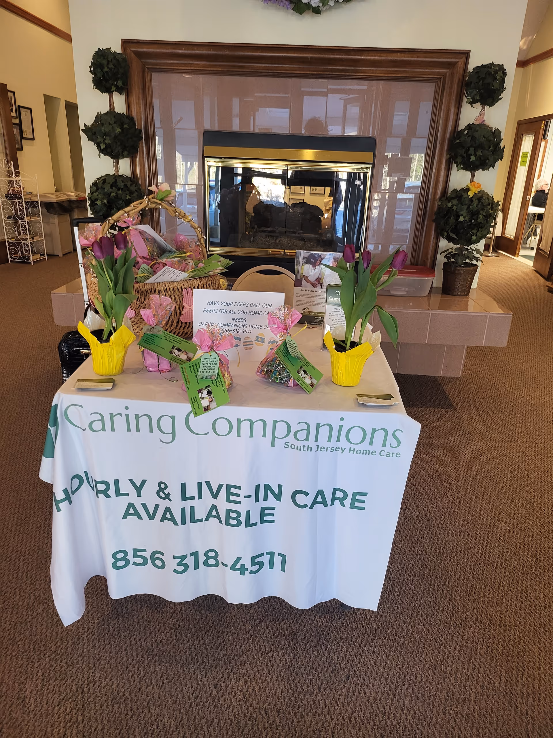 A reception-style table with Caring Companions signage, flowers, and gift baskets in a lobby.