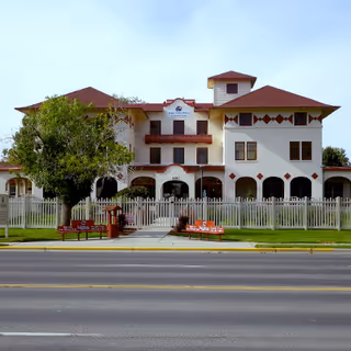 Front view of Magic Valley Manor: a three-story white building with red roofs, arched covered porch, fenced lawn, and benches along the sidewalk.