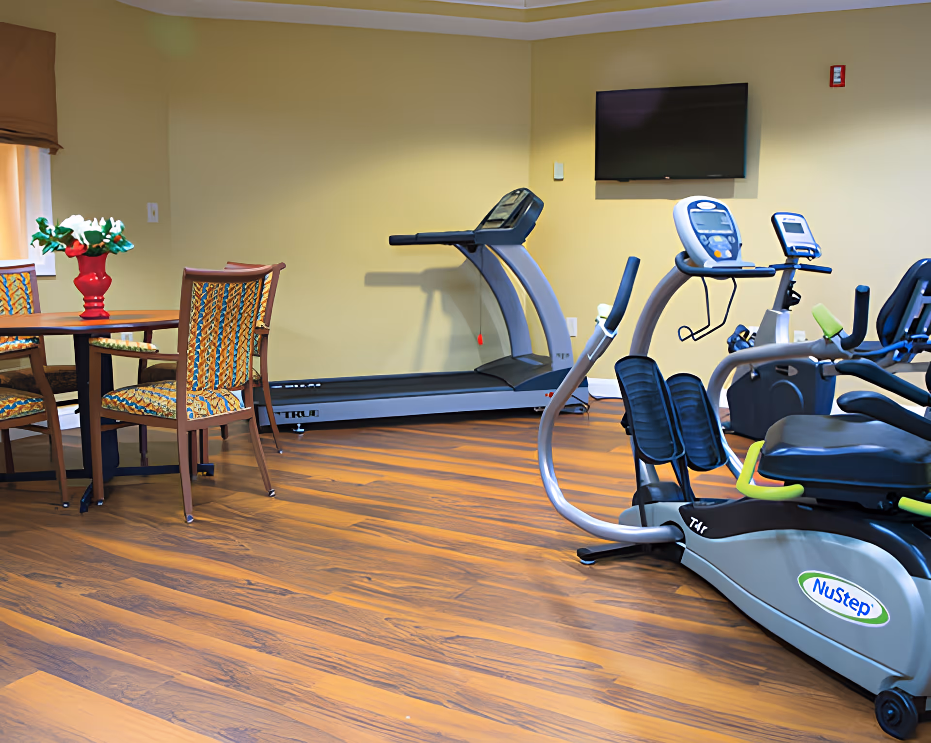 A room with exercise equipment including a treadmill and a NuStep recumbent cross trainer, alongside a round wooden table with four patterned chairs and a red vase with white flowers on it. The room has wooden flooring, yellow walls, and a wall-mounted flat screen TV.