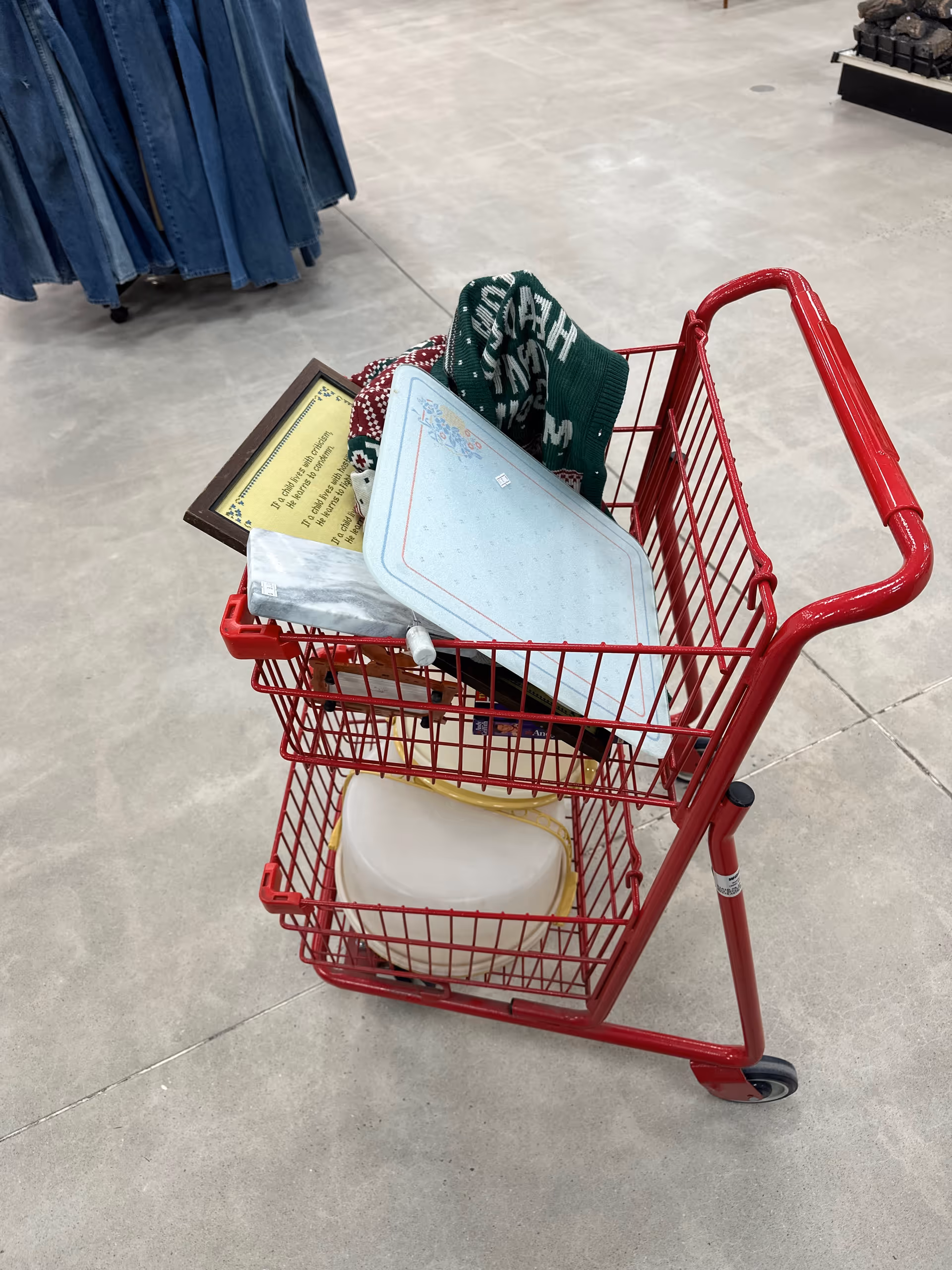 Red shopping cart in a store aisle loaded with blankets, papers, and a white container.