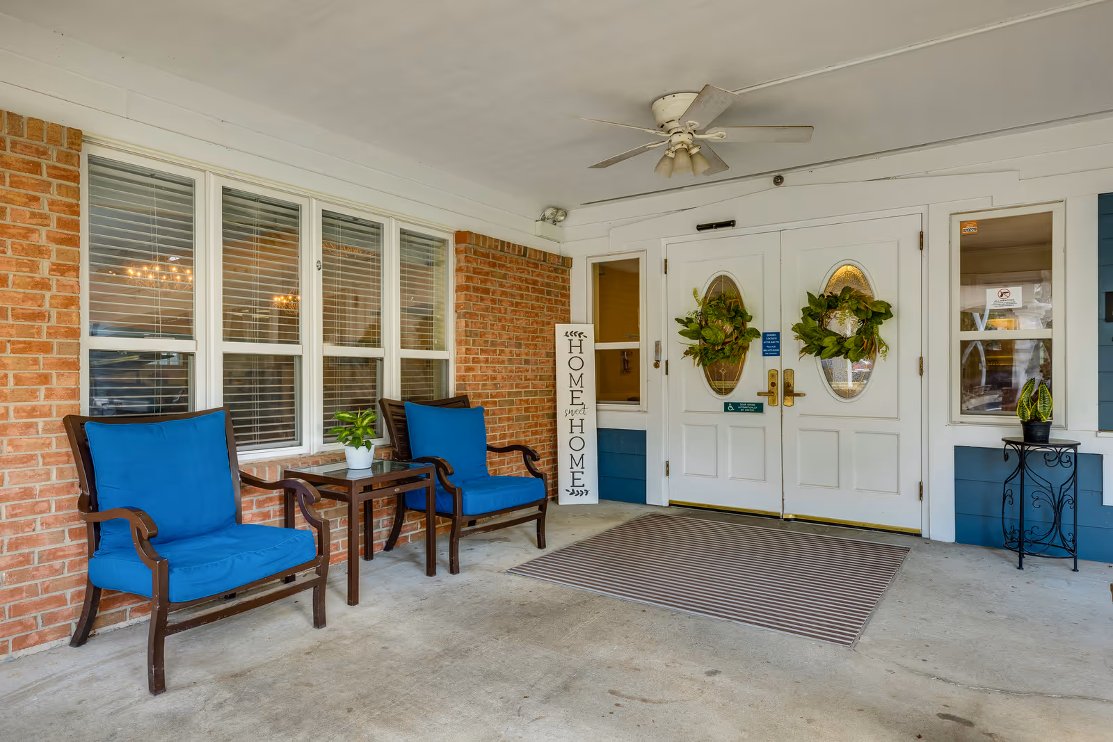 Covered entrance area of a facility with two wooden chairs with blue cushions and a small table with a potted plant between them. The entrance has double white doors decorated with green wreaths, a ceiling fan above, and a vertical sign that reads 'Home sweet Home' leaning against the wall. The walls are a combination of red brick and blue siding with white trim.