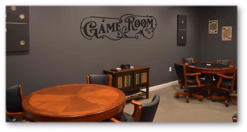 Interior view of a game room with two round wooden tables surrounded by black and wooden chairs. The walls are painted gray and decorated with a 'Game Room' sign and framed artwork resembling playing cards.