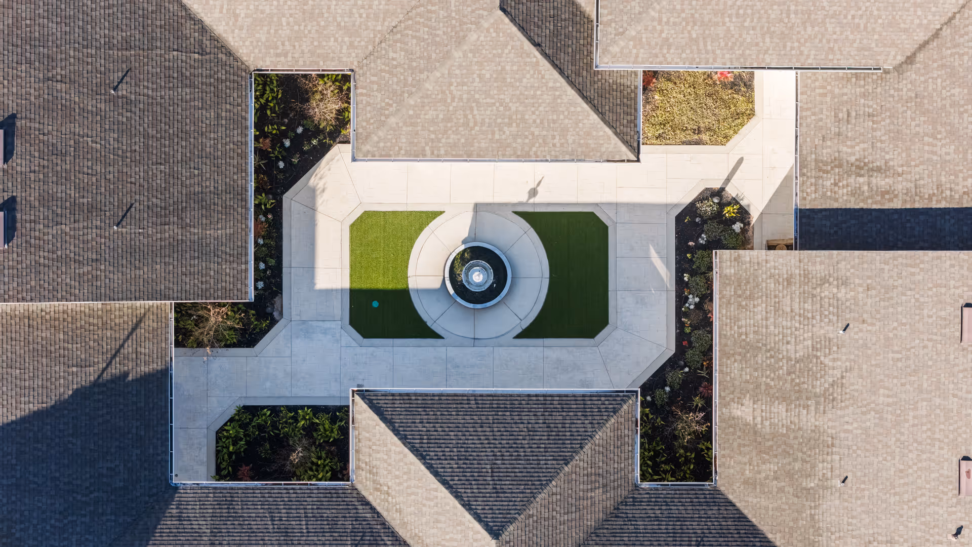 Aerial view of a courtyard area within a building complex, featuring a central circular water fountain surrounded by green artificial turf and concrete walkways. The courtyard is bordered by landscaped garden beds and the roofs of the surrounding buildings.