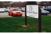 Outdoor scene with grassy lawn, parked cars, and sign for Orchard Rehabilitation and Nursing Center.