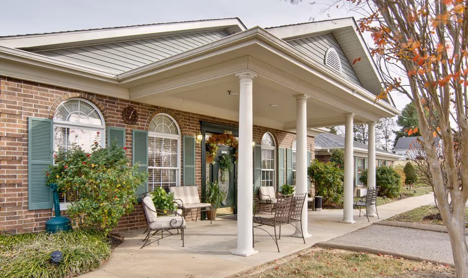 Covered front porch of a brick building with white columns, outdoor seating, arched windows with green shutters, and landscaping.